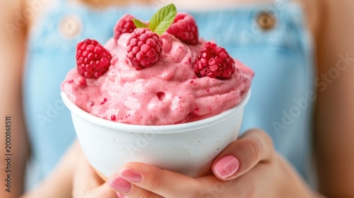 Close up view of a creamy pink raspberry smoothie or ice cream dessert garnished with fresh raspberries and mint held in a white bowl by two hands suggesting a refreshing summer treat