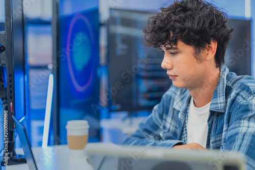 focused young Asian male programmer with curly hair is working intently on his laptop in a modern data center office. The background features bright blue neon LED-lit monitor