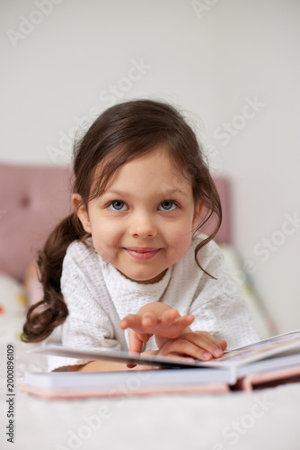 toddler girl with big blue eyes smiling, enjoying a storybook while relaxing in bed, promoting childhood learning