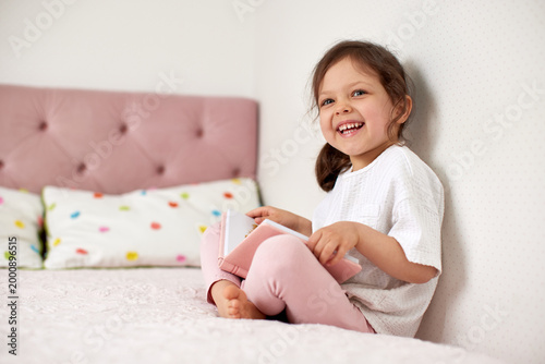 Young girl smiling broadly, holding an open pink book, sitting barefoot on a pastel bed, enjoying childhood at home