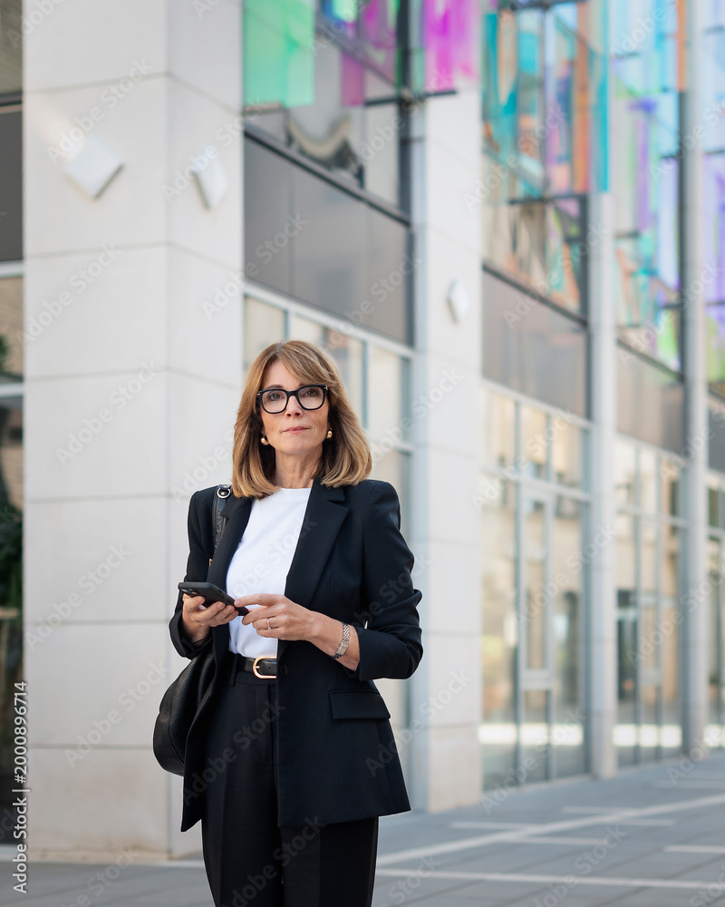 Fototapeta premium Confident businesswoman with smartphone in her hand walking to the office