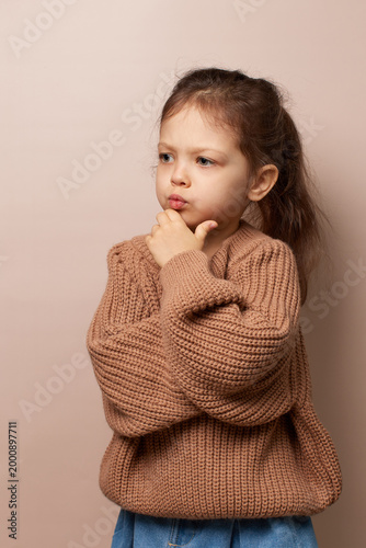 little child girl in a sweater thoughtfully pondering with hand on chin on beige background