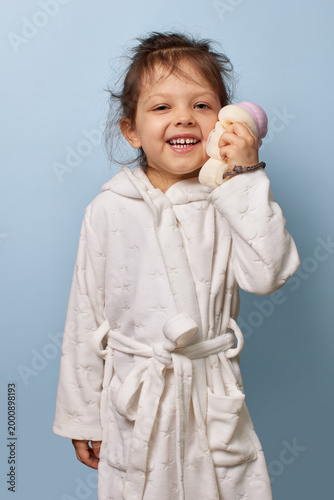 Smiling young girl wearing a white bathrobe and holding a soft bath sponge