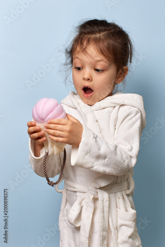 Little child girl in white bathrobe holding and looking at a pink bath sponge with wonder