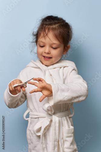 Child girl in a bathrobe learning nail care with scissors on blue background