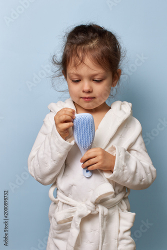 Toddler girl in a bathrobe carefully holding a hairbrush against a blue background