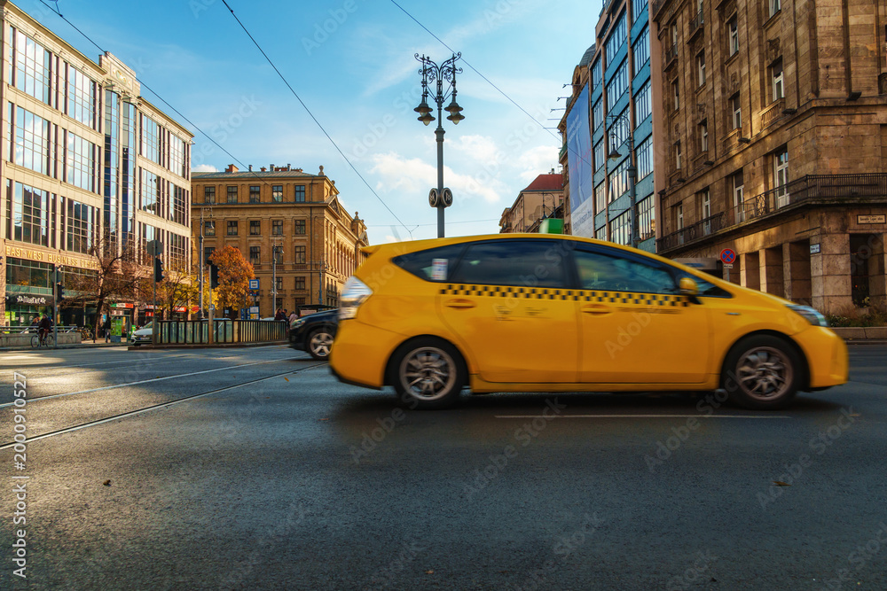 Fototapeta premium Yellow taxi cab driving fast on a city street in Budapest Hungary with blurred motion effect and beautiful European architecture under a bright blue sky during daytime