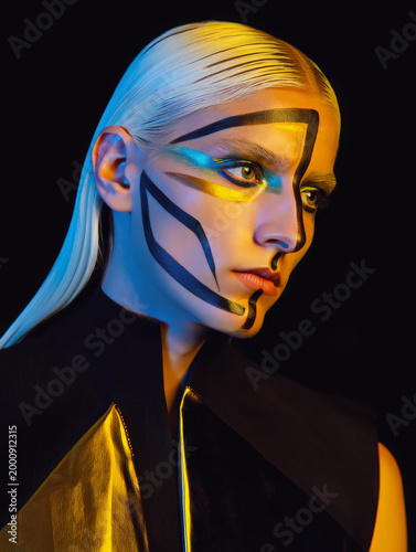 Avant-garde portrait of a woman with slicked-back platinum hair and striking geometric face paint. Bold black lines and metallic gold accents create a futuristic, high-fashion aesthetic