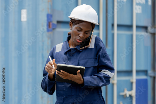 Foto A female worker in a hard hat uses a tablet while talking on the phone at an industrial site with shipping containers