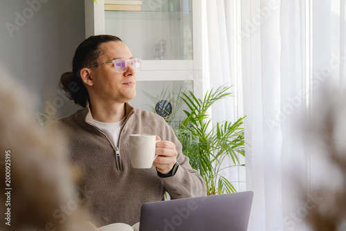 Middle aged man enjoying coffee break while working remotely near bright window. Peaceful pause, work life balance, productivity and calm home office routine concept.