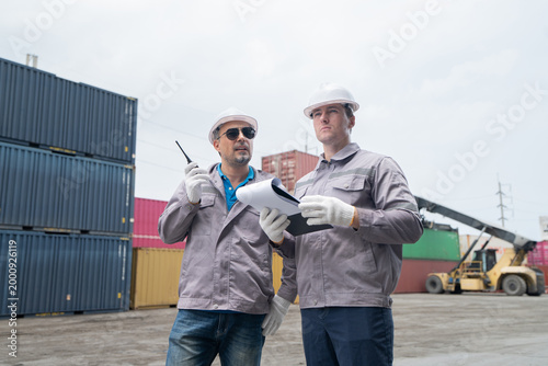 Two workers in safety gear inspect shipping containers at an industrial site, discussing logistics with documents and a walkie-talkie.