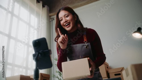 Young asian female business owner live streaming on her smartphone, presenting a product in a box for sale to her online customers