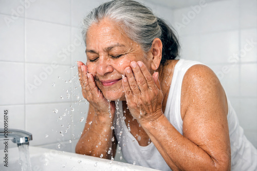 Senior woman splashing water on face at bathroom sink.