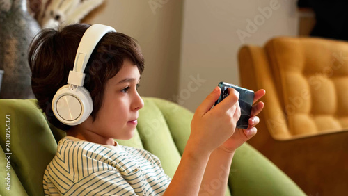 Side view of a boy wearing white headphones while holding a smartphone on a green sofa at home. Modern childhood, screen time, mobile games, and digital habits shape family life and leisure.