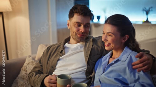 Happy lovers embracing sofa drinking evening coffee together. Man cuddling woman