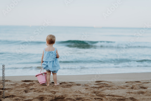 Bild auf Leinwand Toddler standing on sand beach with bucket in hand.