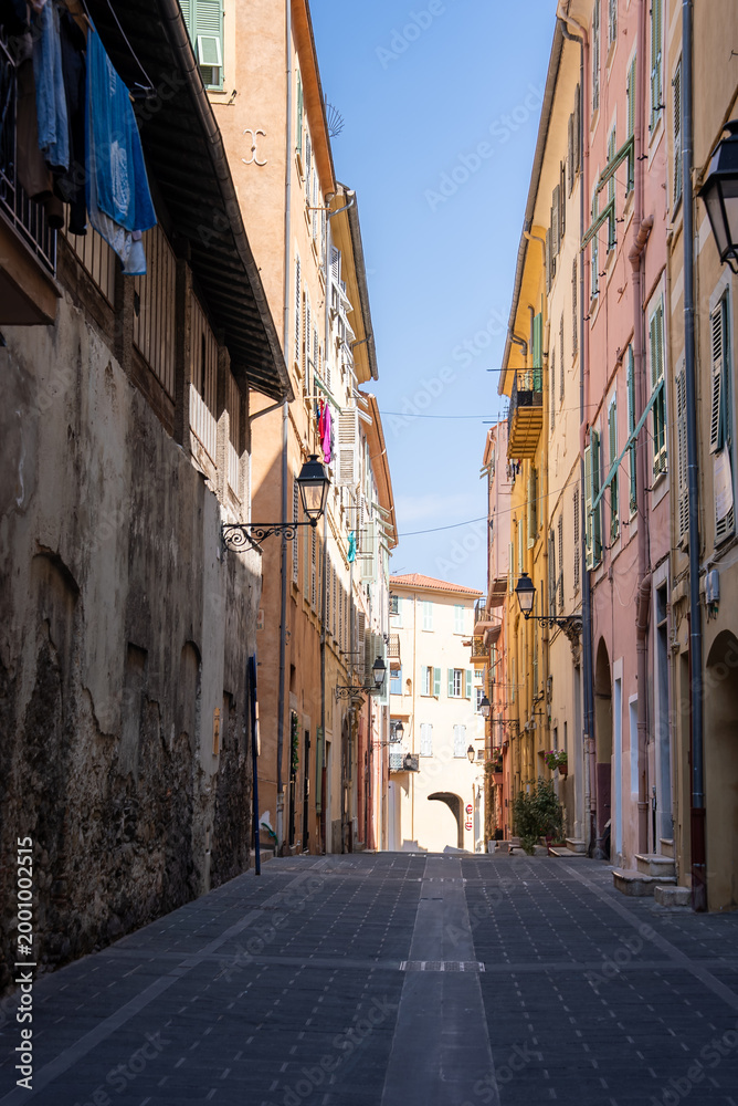 Fototapeta premium A narrow alley in Menton, France, leads to an archway. Pastel buildings with green shutters, wrought iron lamps, and laundry hang in crisp midday light and deep shadows.