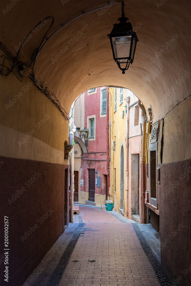 Obraz premium A narrow vaulted passage opens to a quiet alley in Old Town Menton, France. Pastel facades, a wrought iron lantern, shutters, a small balcony, and a blue bucket stand out.