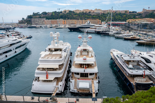 Luxury yachts line Port Hercule in Monaco, with Le Rocher and the Prince's Palace across the water. Daylight scene shows glossy decks, masts, cranes, and calm weather.