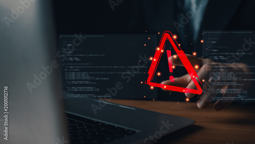 Programmer pointing at a red virtual warning triangle icon on a screen with coding background. Concept of cyber attack, system error, data breach, software bug, and internet security alert.