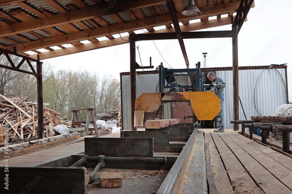 Fototapeta premium A worker controls a log sawing machine. Production of wooden pallets for heavy cargo transportation. An open-air locksmith workshop. The concept of woodworking.