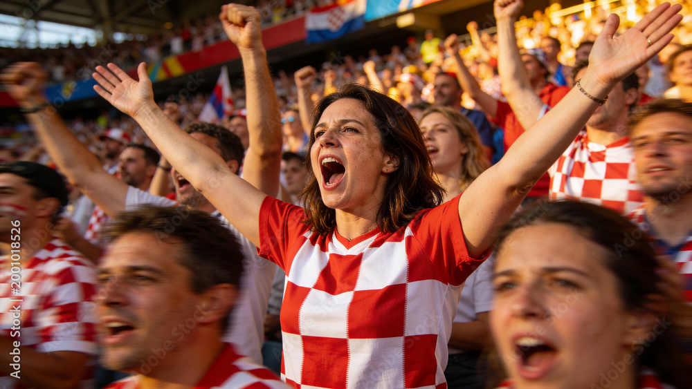 Fototapeta premium Cheering female fan raising arms in stadium stands, wearing red-and-white checkerboard jersey