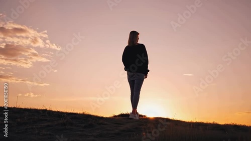Adult woman with blonde hair stands on hilltop, gazing at sunset over horizon, surrounded by grass and open sky, capturing serene moment in nature