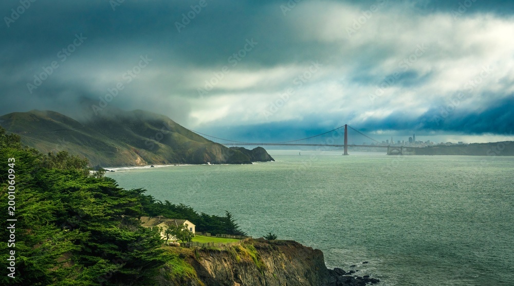 Naklejka premium Golden Gate Bridge partially visible through coastal fog with cliffs and ocean viewed from a scenic hillside in natural light.