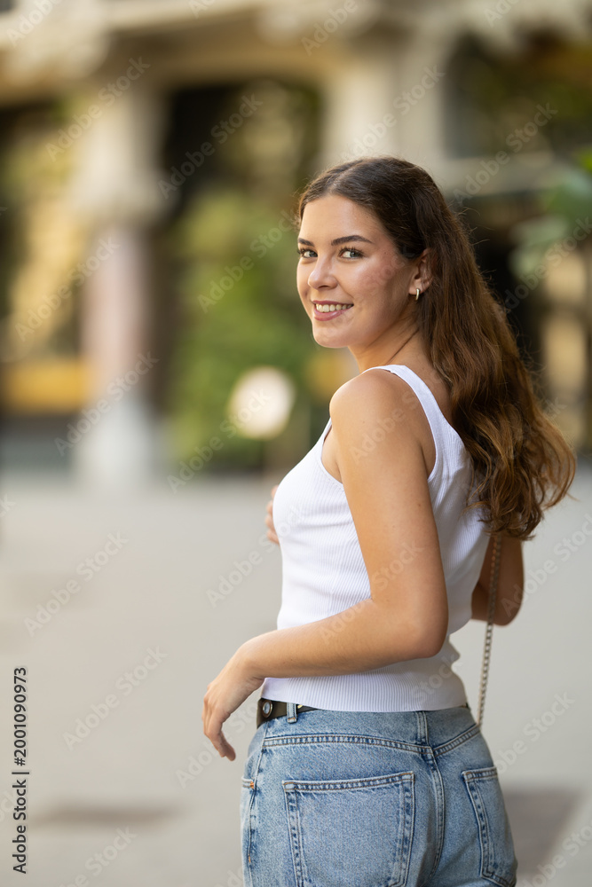 Fototapeta premium Young smiling girl walks along city street, walks from work to home. Pedestrian in summer European capital, person in city, city bustle.
