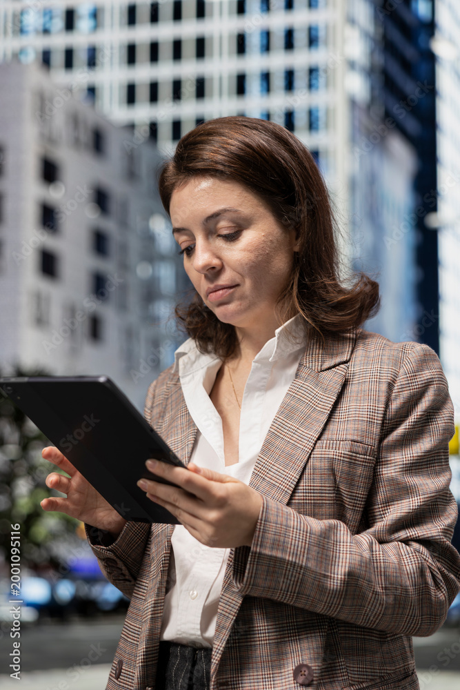 Fototapeta premium Businesswoman using tablet to browse online content, walking on city boulevard. Person using touchscreen device, relaxing by looking at social media posts on feed, urban cityscape background