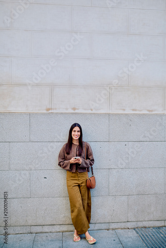 Young woman smiling, standing outdoors by a wall, wearing a brown cardigan and brown trousers, using a mobile phone