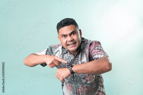 Tableau sur toile An angry Asian man in a batik shirt pointing at his watch with a furious expression, representing lateness, deadlines, and time management stress