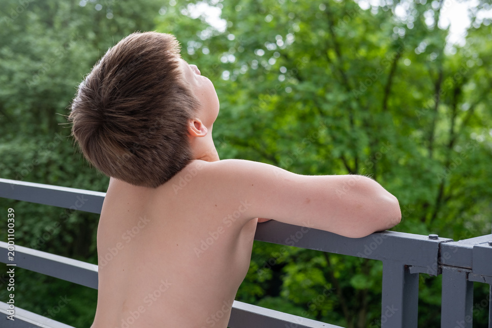 Naklejka premium Young Shirtless boy standing on terrace, balcony, enjoying fresh air outdoors, summer day, peaceful Back view, green forest background, childhood moment, nature connection