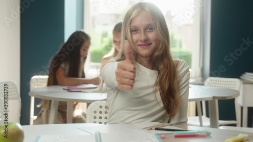 Happy Caucasian little girl child showing thumb up studying in classroom recommend gesture kid schoolgirl student pupil teenager writing lesson task exam success education looking camera approve like