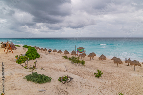 Принти на полотні A wide view of a white sand beach in Cancun featuring rows of straw palapa umbrellas and a colorful lifeguard tower under a cloudy sky