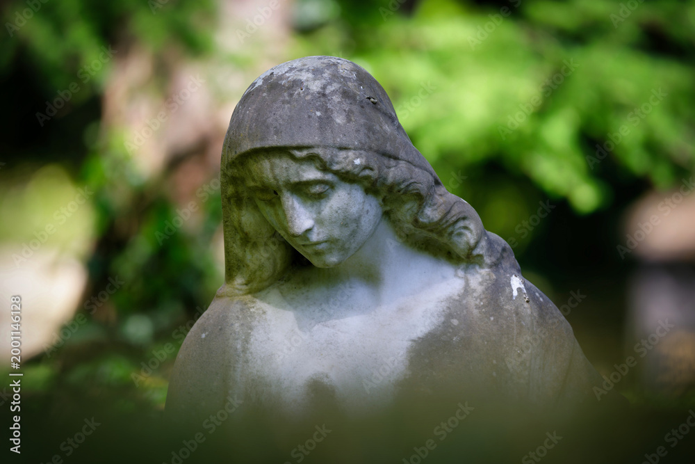 Naklejka premium Close-up of a melancholy stone female statue with a downward gaze against a softly blurred green natural background