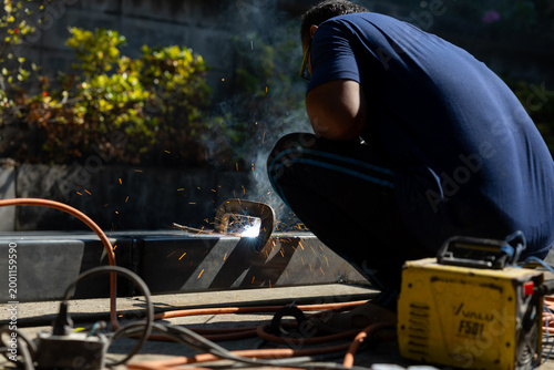 Welder in blue shirt squatting while welding metal with bright sparks and smoke