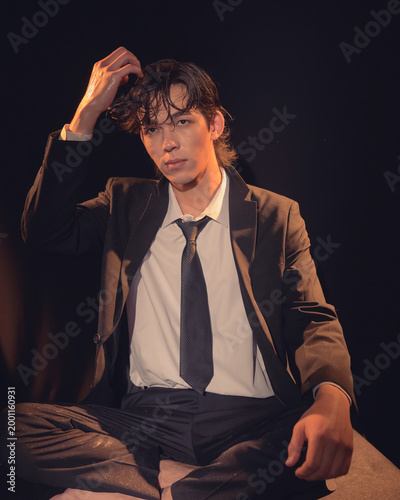 Φωτογραφία The image portrays a young man in a stylish suit, casually adjusting his hair while seated against a dark background, showcasing a relaxed yet confident demeanor enhanced by rain effects