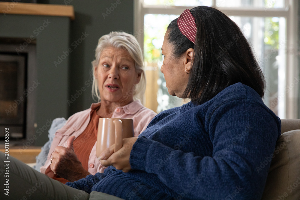 Fototapeta premium Senior female friends chatting while holding ceramic mugs on living room sofa near fireplace