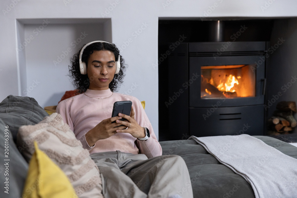 Obraz premium Non-binary person lying on gray sofa at home, wearing white headset and checking phone