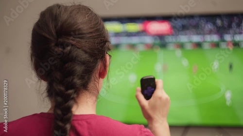 Frustrated young Mexican woman watching a soccer match on television, using a remote control and showing signs of disappointment with the game's outcome from her living room