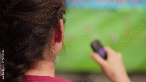 Woman with braided hair using a remote control to change channels while watching a soccer cup on a big screen television, enjoying her free time in a modern living room
