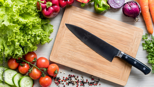 A top-down overhead shot of a professional chef knife on a wooden cutting board surrounded by vibrant fresh vegetables like lettuce, tomatoes, and carrots.
