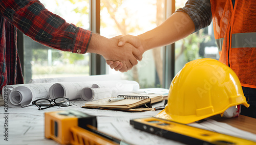 Two construction workers shaking hands over blueprints and safety gear on a desk.