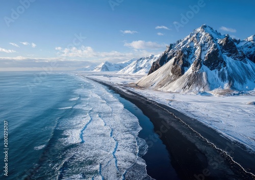 A breathtaking aerial view showcases a dramatic black sand beach meeting the turquoise ocean, framed by snow-capped mountains under a clear blue sky, creating a stunning landscape.