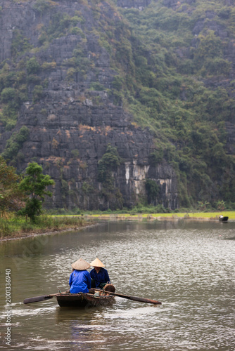 Traditional boat with two rowers wearing conical hats sailing on the river in Tam Coc, Ninh Binh region, Vietnam.