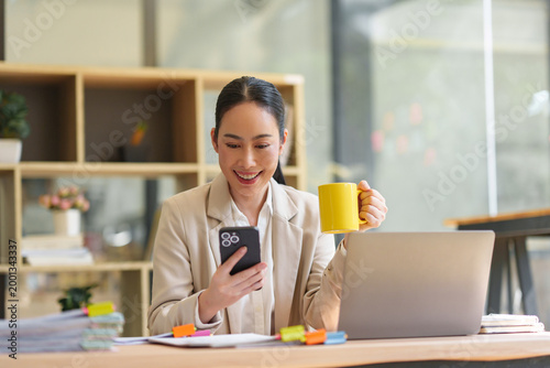 Young Asian businesswoman smiling while checking social media on smartphone, holding a yellow mug, and working with a laptop in office