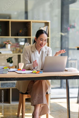 Professional Asian woman smiling, engaged in a virtual meeting on her laptop at an office desk, communicating and collaborating online