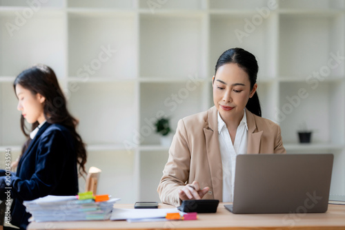 Two businesswomen working at their desks, one focused on calculating finances with a calculator and laptop, the other working in background