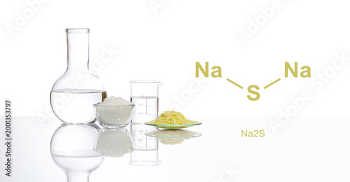 Sodium sulfide flake in Chemical Watch Glass place next to beaker on white laboratory table. The structure of a chemical compound is shown on the side.